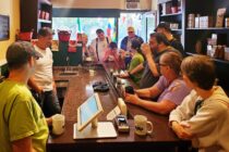 Ten people gather around the long wooden counter at 9th Street Coffee & Tea. Jake, behind the counter in a white shirt, tells the group about his company.